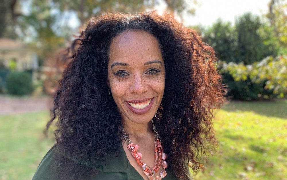 A woman with long, curly hair smiles outdoors with sunlit trees behind her. She is wearing a green top and a pink necklace.